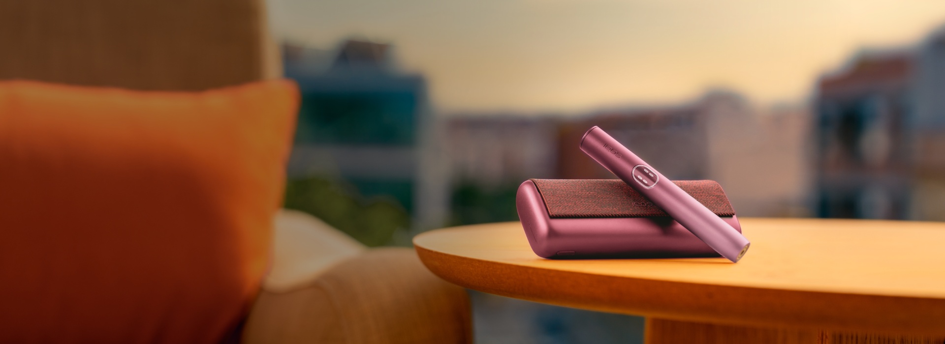 Stylish Garnet Red IQOS ILUMA i PRIME on a wooden table with a blurred cityscape background, next to a cozy armchair with an orange cushion.